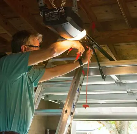 A person repairing a garage door opener from a ladder.